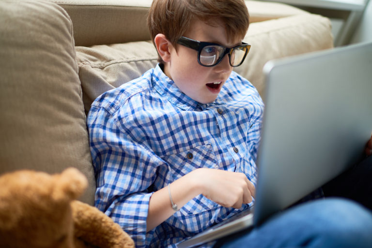 Shocked boy in eyeglasses sitting on sofa and using laptop, he playing video game